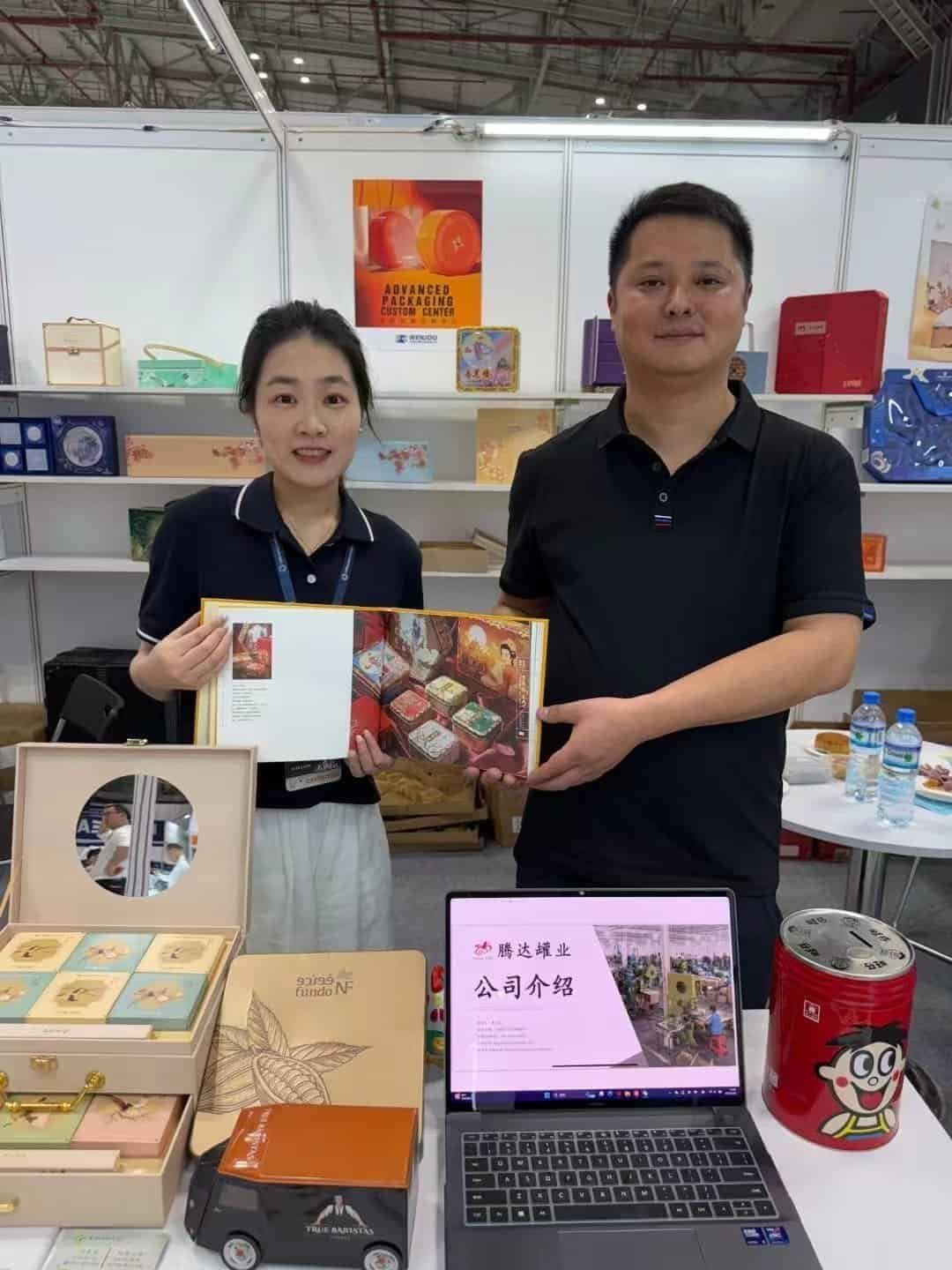 Two people holding a printed tin box in front of a display of tin packaging samples